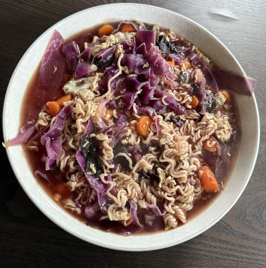 A bowl of pho-style ramen with purple cabbage, carrots, shredded egg, and noodles in a rich broth.